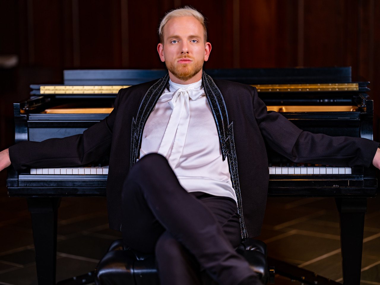 Stephen Aber seated in front of a black grand piano, arms outstretched along the keyboard, wearing a white bow blouse and black embellished jacket